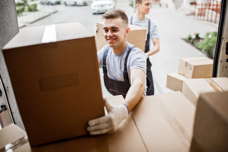 A young handsome smiling mover wearing uniform is reaching for the box while unloading the van full of boxes. House move, mover service.